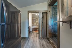 Ein schmaler Flur mit einem Edelstahl-Kühlschrank links, einem Holzrahmen einer Tür rechts und einem durch eine offene Tür einsehbaren Badezimmer in der Mitte.