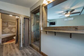 Ein Badezimmer mit Dusche, Toilette und Waschbecken. An der Wand befindet sich ein Holzregal und ein Deckenventilator.