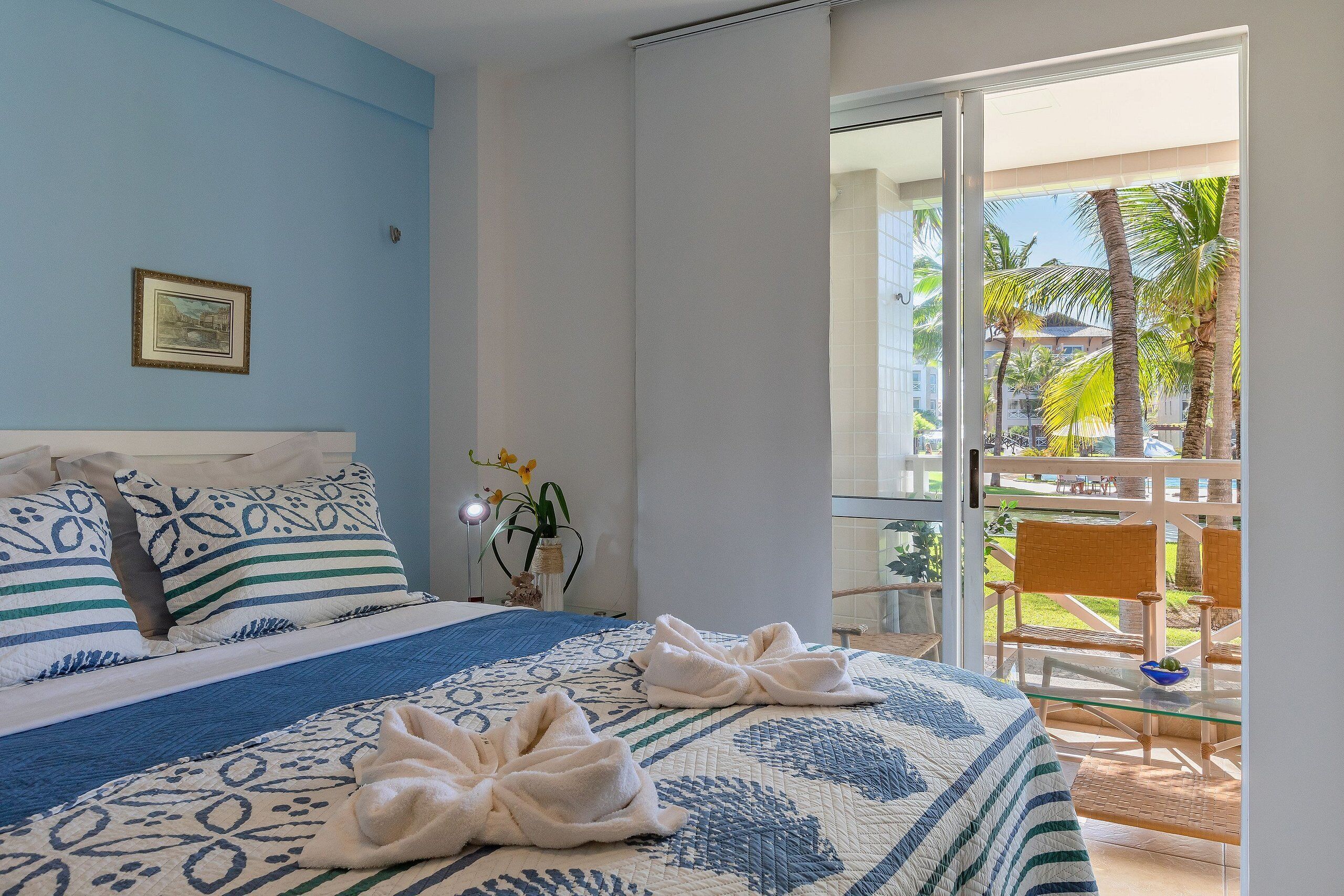 Una habitación de hotel con cama, un balcón con sillas y vista a palmeras.