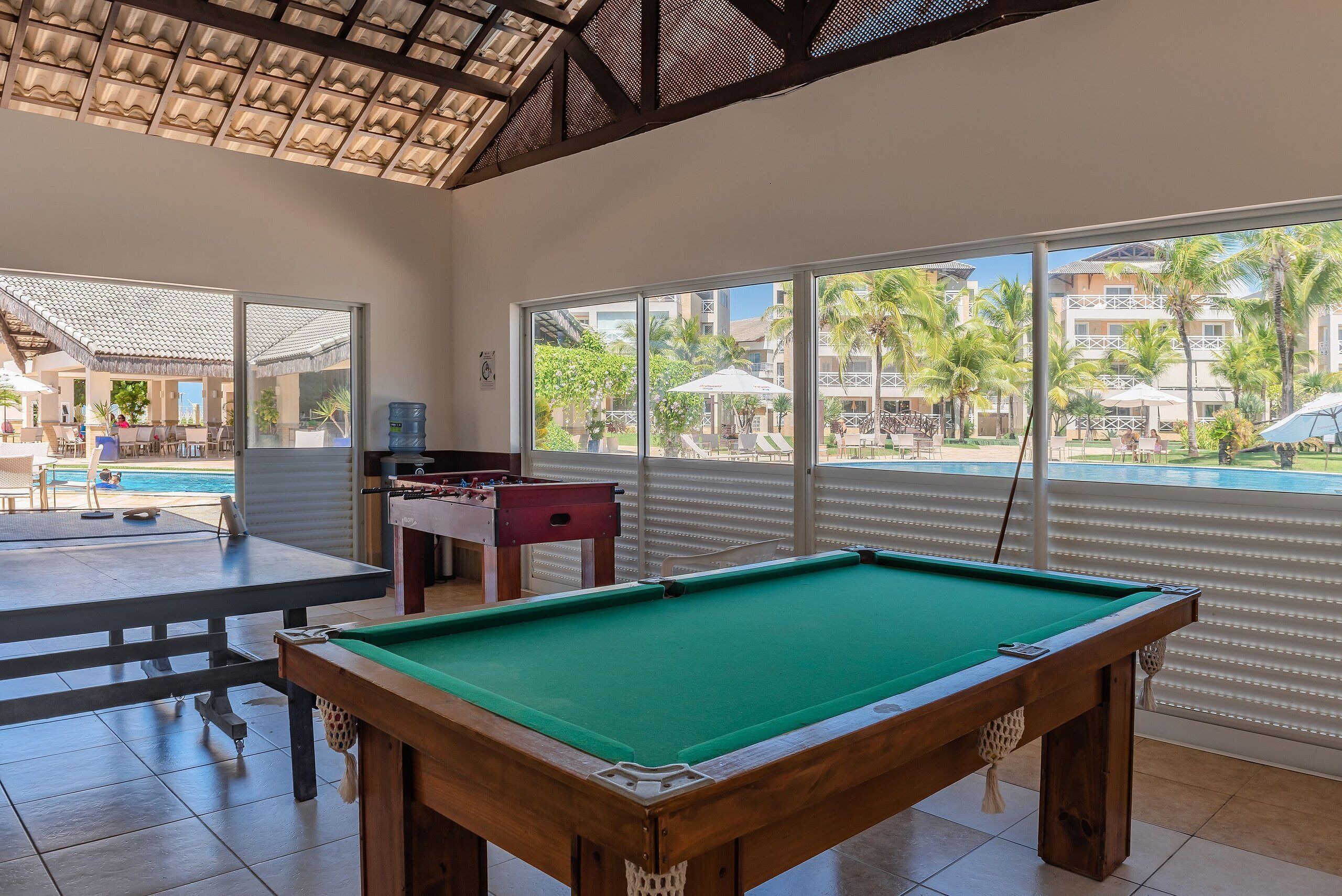 Mesa de billar y mesa de futbolín en una habitación con ventanas amplias que dan a una piscina y zona de resort.