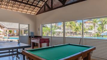 Mesa de billar y mesa de futbolín en una habitación con ventanas amplias que dan a una piscina y zona de resort.