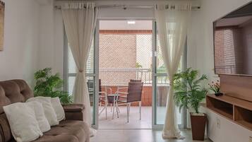 Una sala de estar con sofá, televisor y puerta corrediza de vidrio que da a un balcón con mesa y sillas.