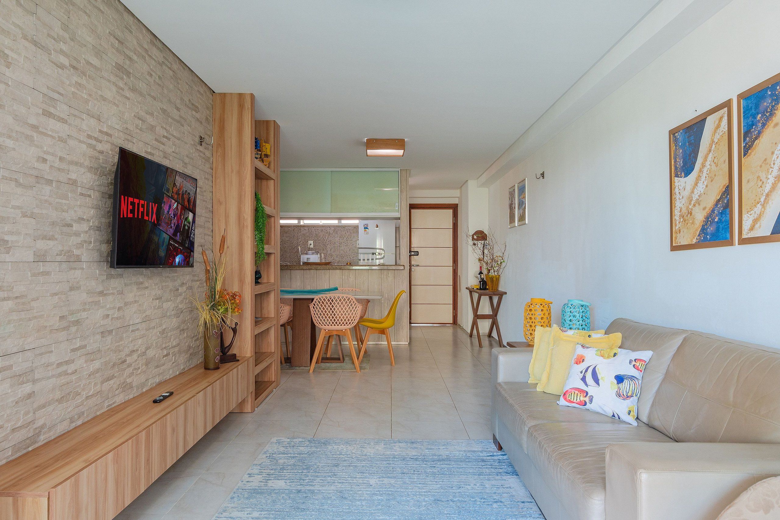 Una sala de estar moderna con un televisor de pantalla plana, un sofá con almohadones coloridos, un mueble bajo de madera y un comedor con mesa y sillas.
