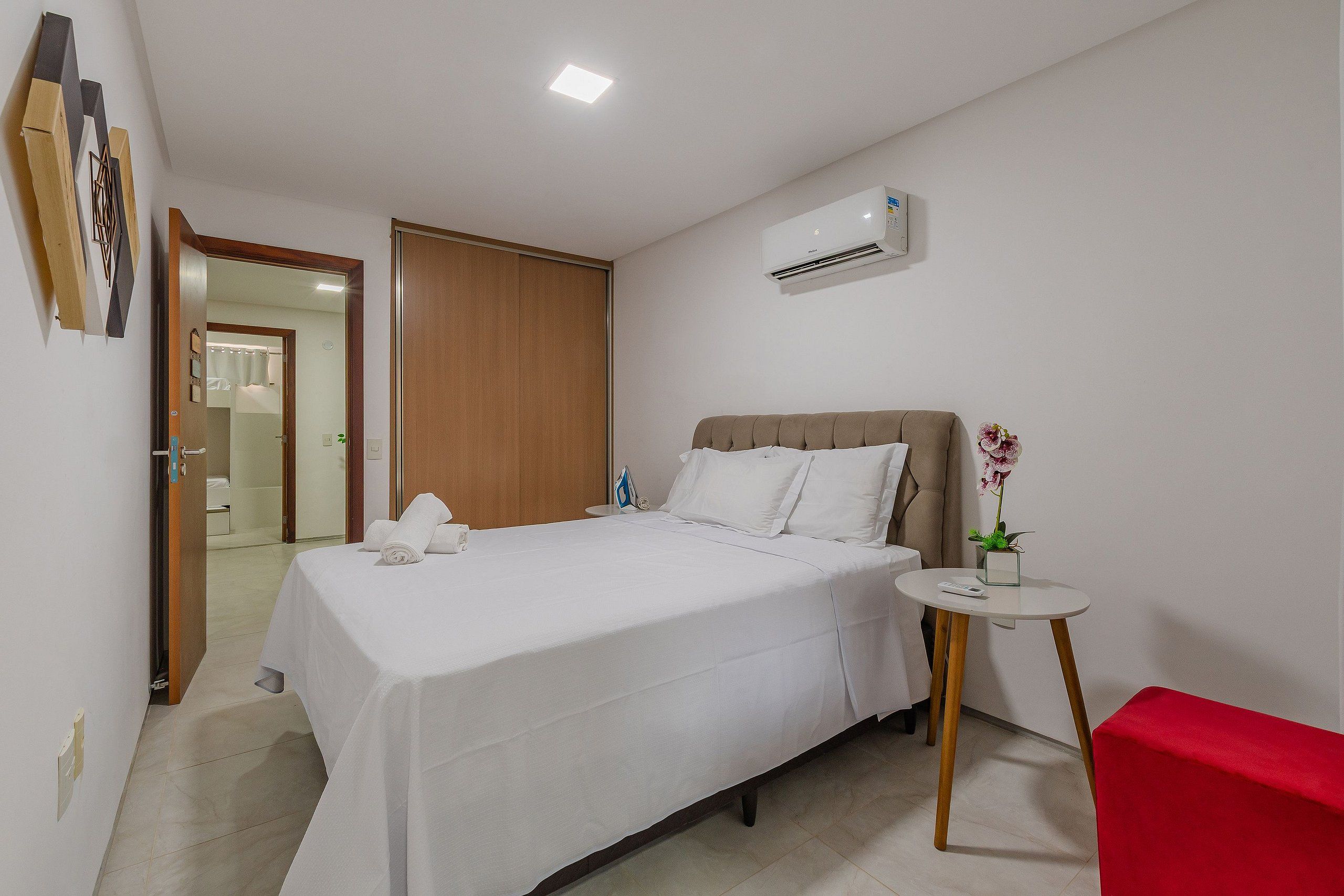 Quarto de hotel com cama, uma mesinha com um vaso de flores, uma cadeira vermelha e um aparelho de ar condicionado na parede.