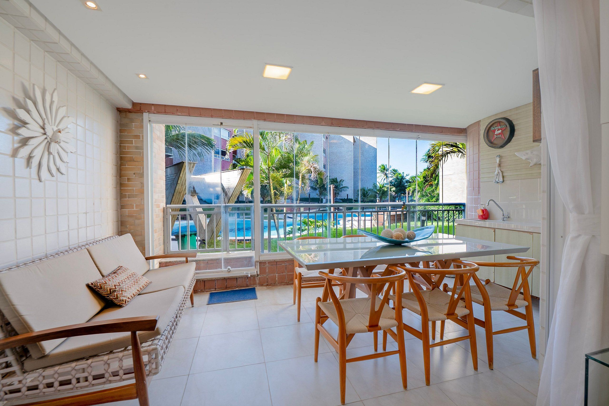 A modern living room with a glass table, wooden chairs, a sofa, and a view of a pool and palm trees.