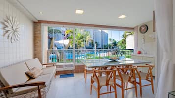 Una sala moderna con una mesa de vidrio, sillas de madera, un sofá y vistas a una piscina y palmeras.