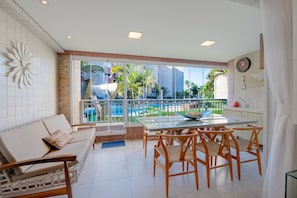 Una sala moderna con una mesa de vidrio, sillas de madera, un sofá y vistas a una piscina y palmeras.