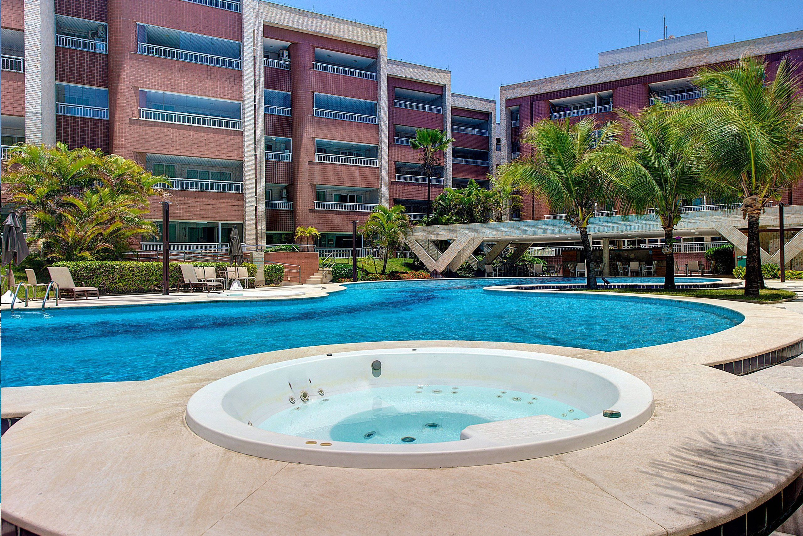 Una piscina con hidromasaje, palmeras y un edificio de varios pisos con balcones.