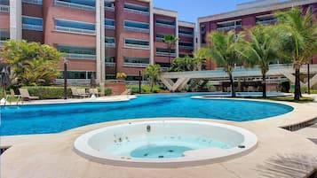Una piscina con hidromasaje, palmeras y un edificio de varios pisos con balcones.