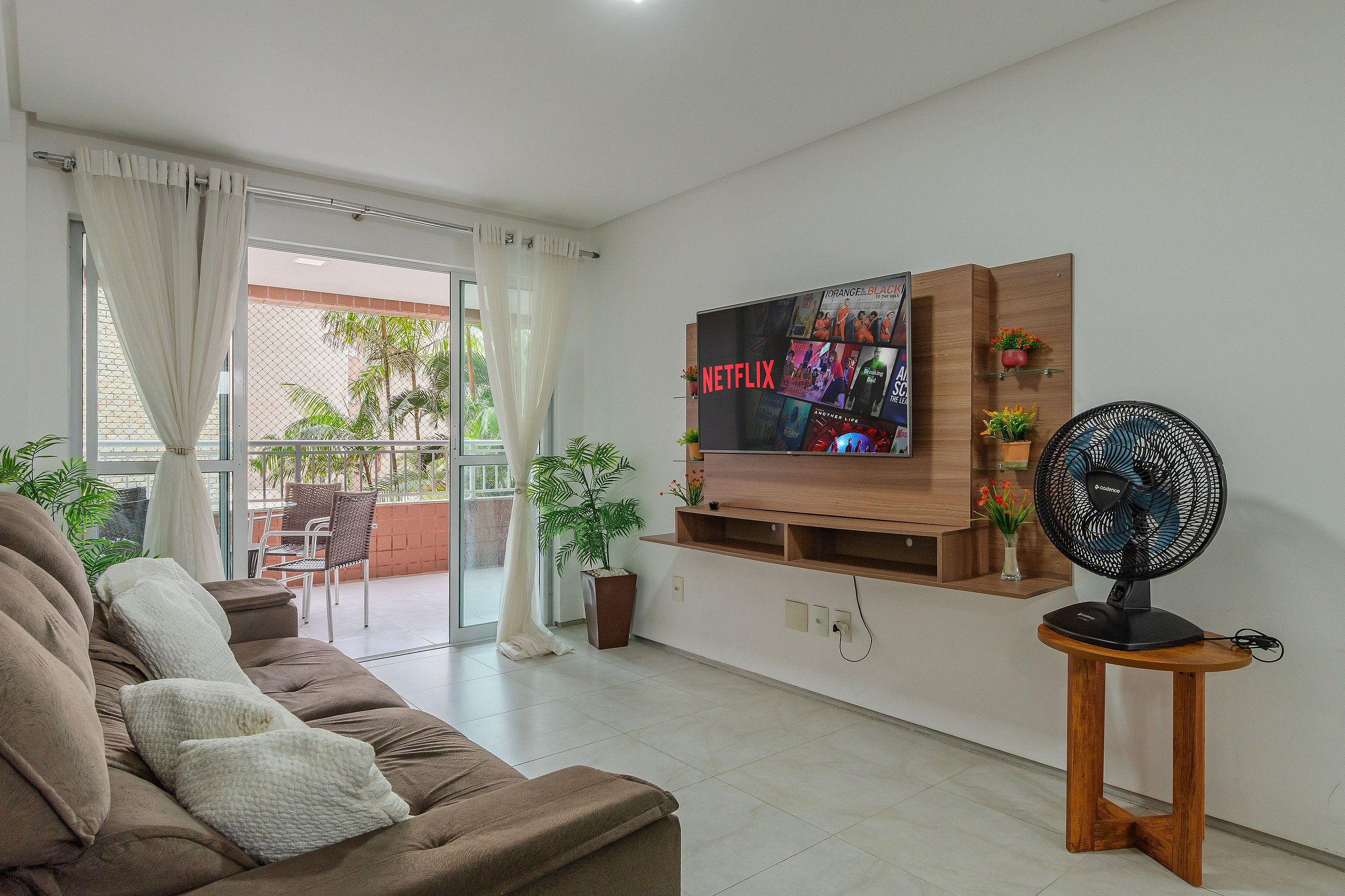 A living room with a sofa, a TV mounted on the wall, a fan, and a balcony with plants.