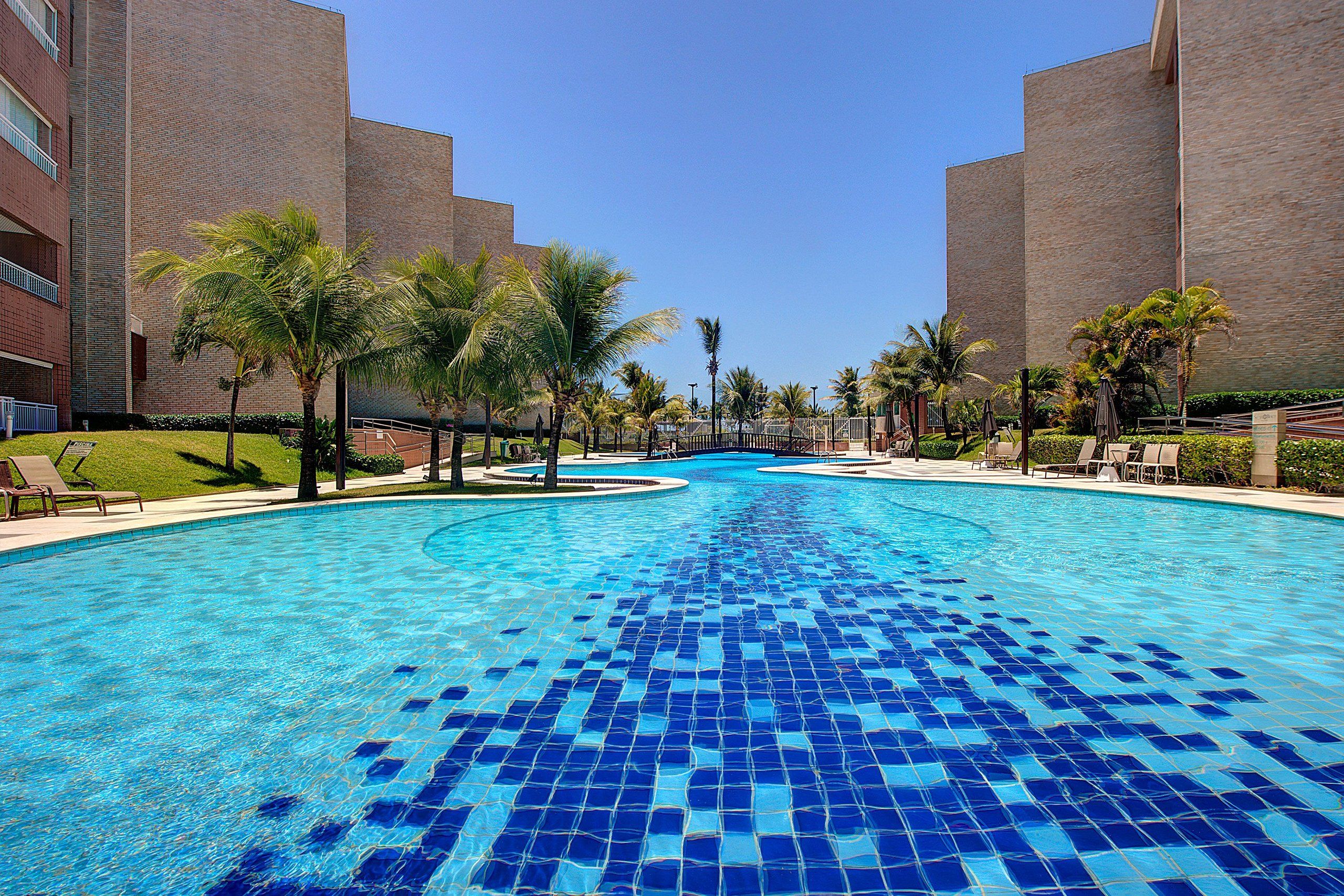 Una piscina con mosaicos azules, rodeada de palmeras y un edificio de ladrillo.