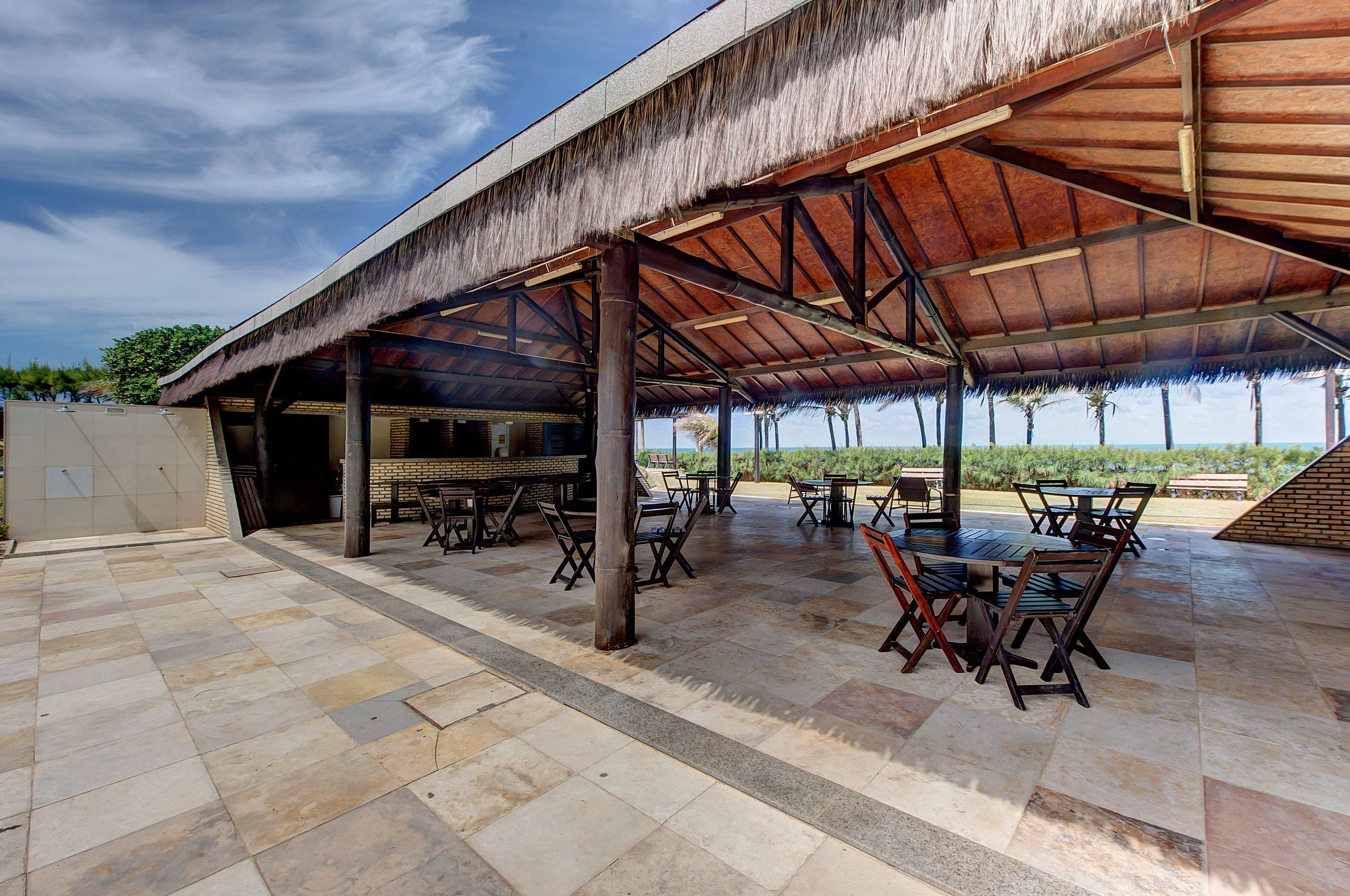 Un patio exterior con techo de paja, suelo de piedra y mesas y sillas de madera.