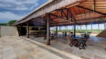 Un patio exterior con techo de paja, suelo de piedra y mesas y sillas de madera.