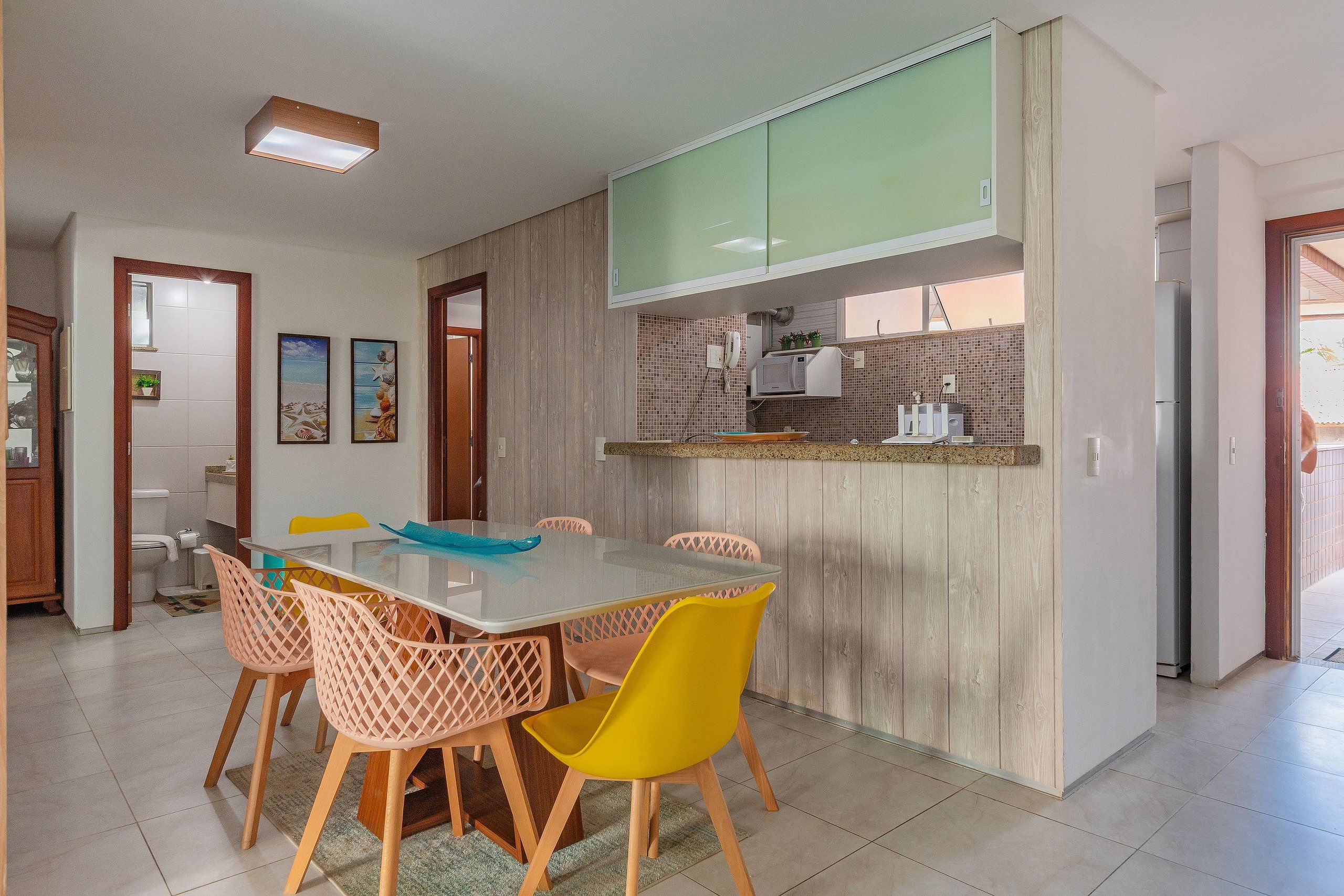 Uma cozinha moderna com micro-ondas embutido, um espaço de refeições com mesa de vidro e cadeiras coloridas, e um armário de madeira sobre o balcão.