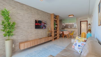 Una sala moderna con televisor de pantalla plana, mueble de madera para entretenimiento, sofá con cojines estampados, comedor con mesa y sillas, y una cocina al fondo.