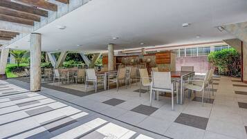 Un área de comedor al aire libre con mesas y sillas, un espacio de estar cubierto y vistas al interior del edificio.