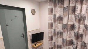 Ein Wohnzimmer mit grauer Tür, einer Wanduhr, einem Flachbildfernseher auf einem Holzregal, einem gemusterten Vorhang und einem kleinen Blumentopf auf einem Couchtisch.