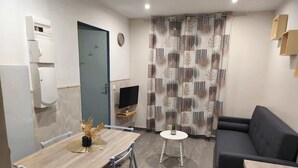 Ein kompakter Wohnraum mit Esstisch, Sofa, Fernseher und einem gemusterten Vorhang.