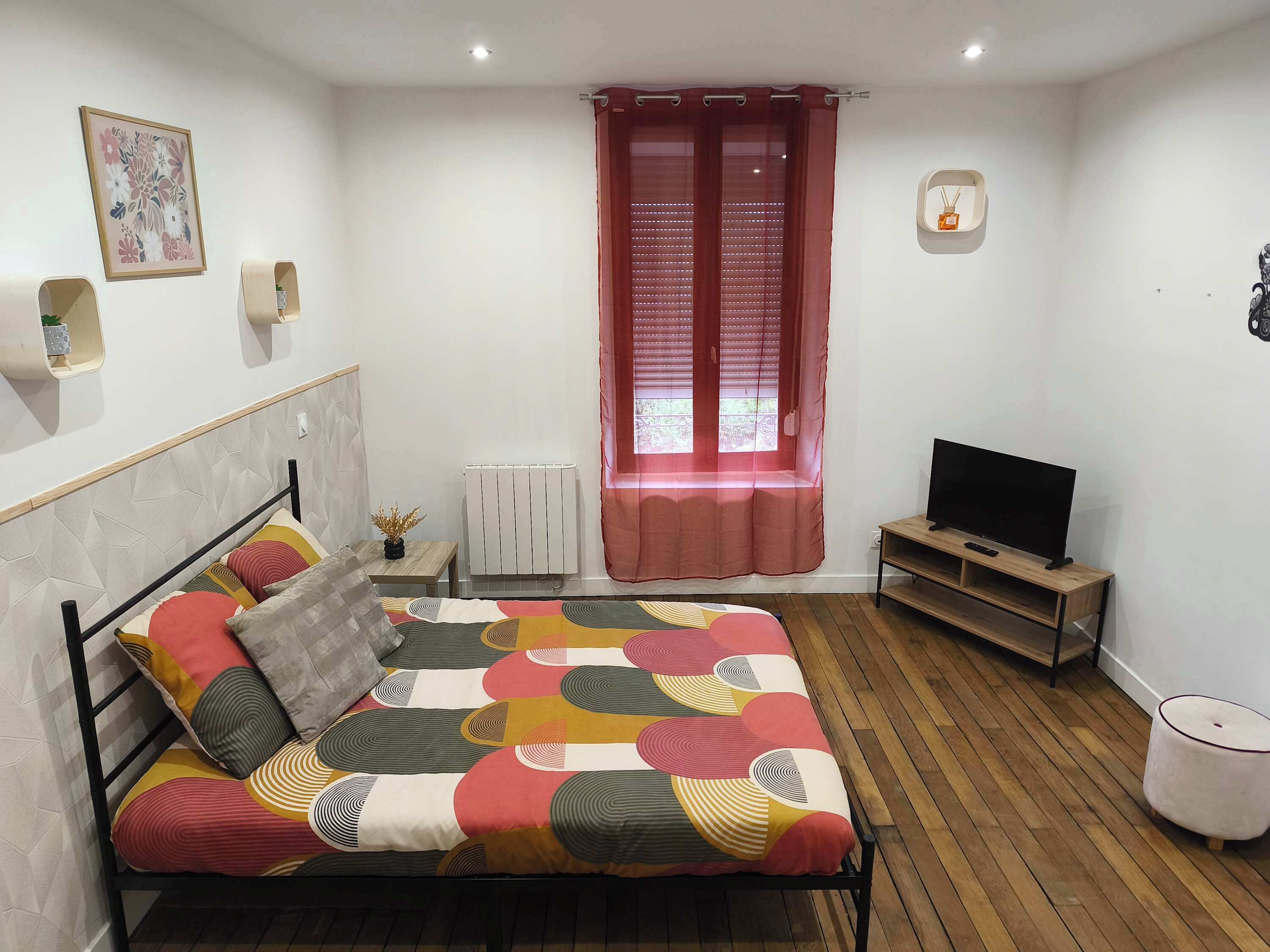 Een slaapkamer met een bed, een televisie, een radiator, een raam met gordijnen en een wandplank.