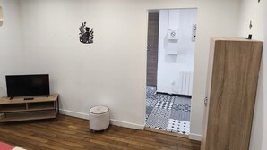 Ein Zimmer mit Holzboden, einem Fernseher auf einem Holzgestell, einer weißen Wand mit einer schwarz-weißen Tür und einem kleinen weißen Hocker.
