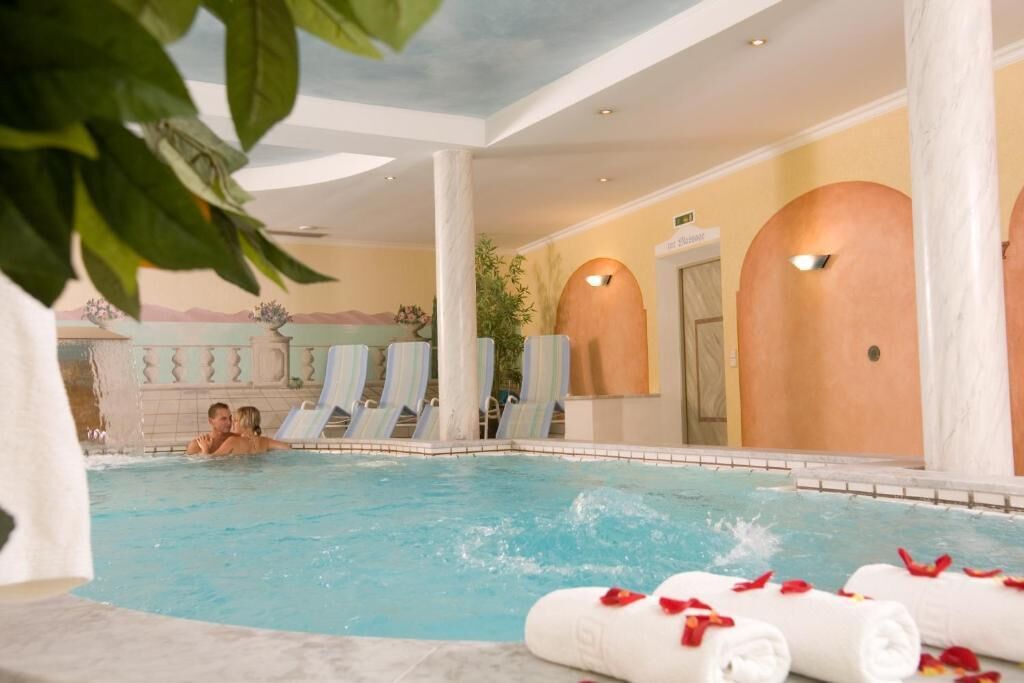 Zona de piscina con sillones de descanso, una pareja en el agua y toallas con pétalos de rosa.