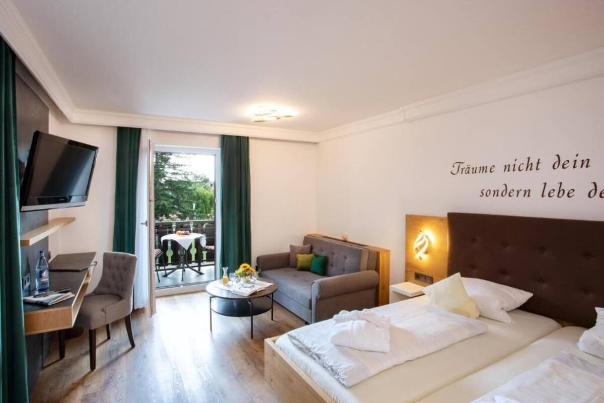 Habitación de hotel con cama, sofá, televisión, una mesita y balcón con mesa y sillas.