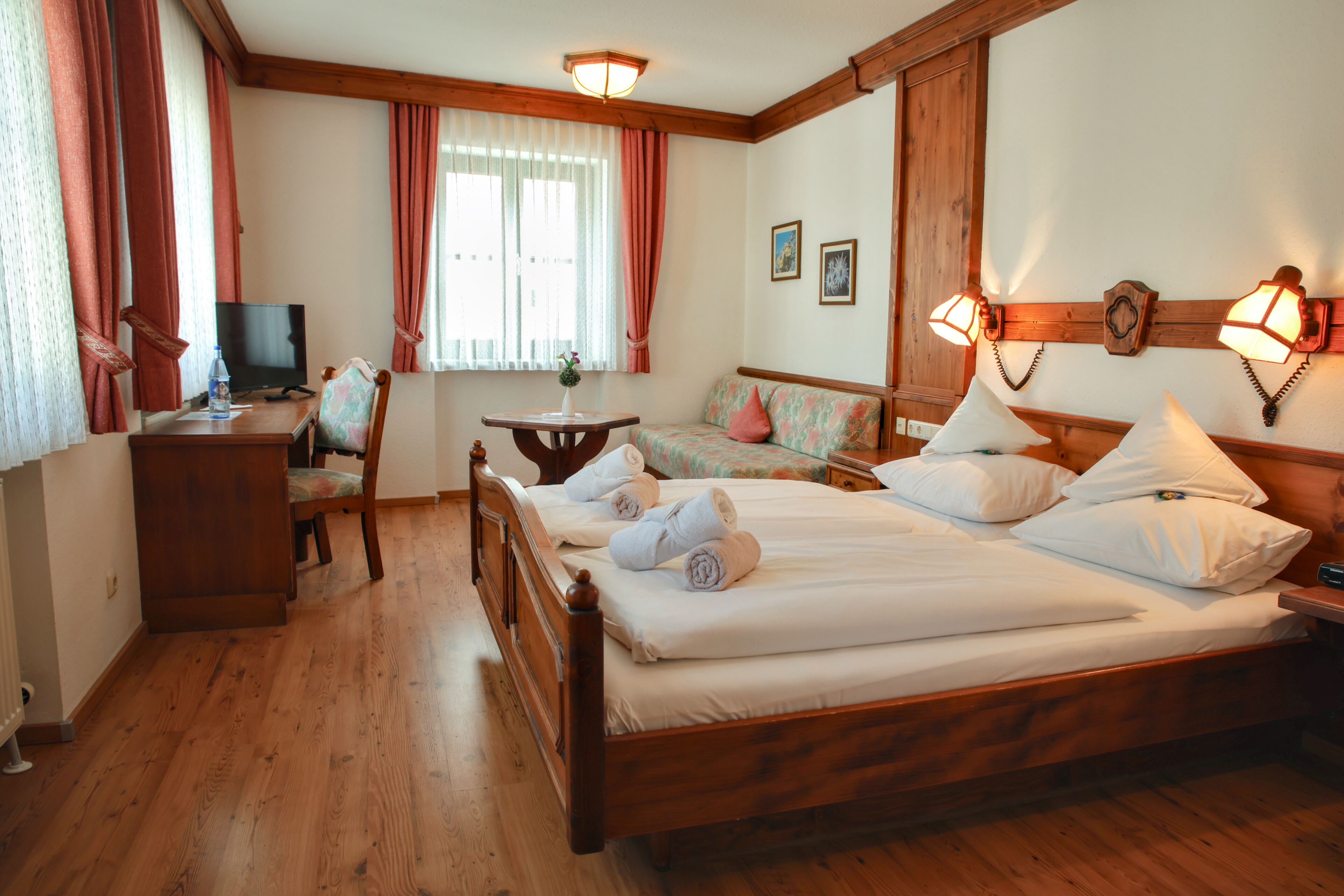 Une chambre d’hôtel avec un lit en bois, un bureau avec une télévision, une chaise, une petite table avec un vase et une fenêtre avec des rideaux.