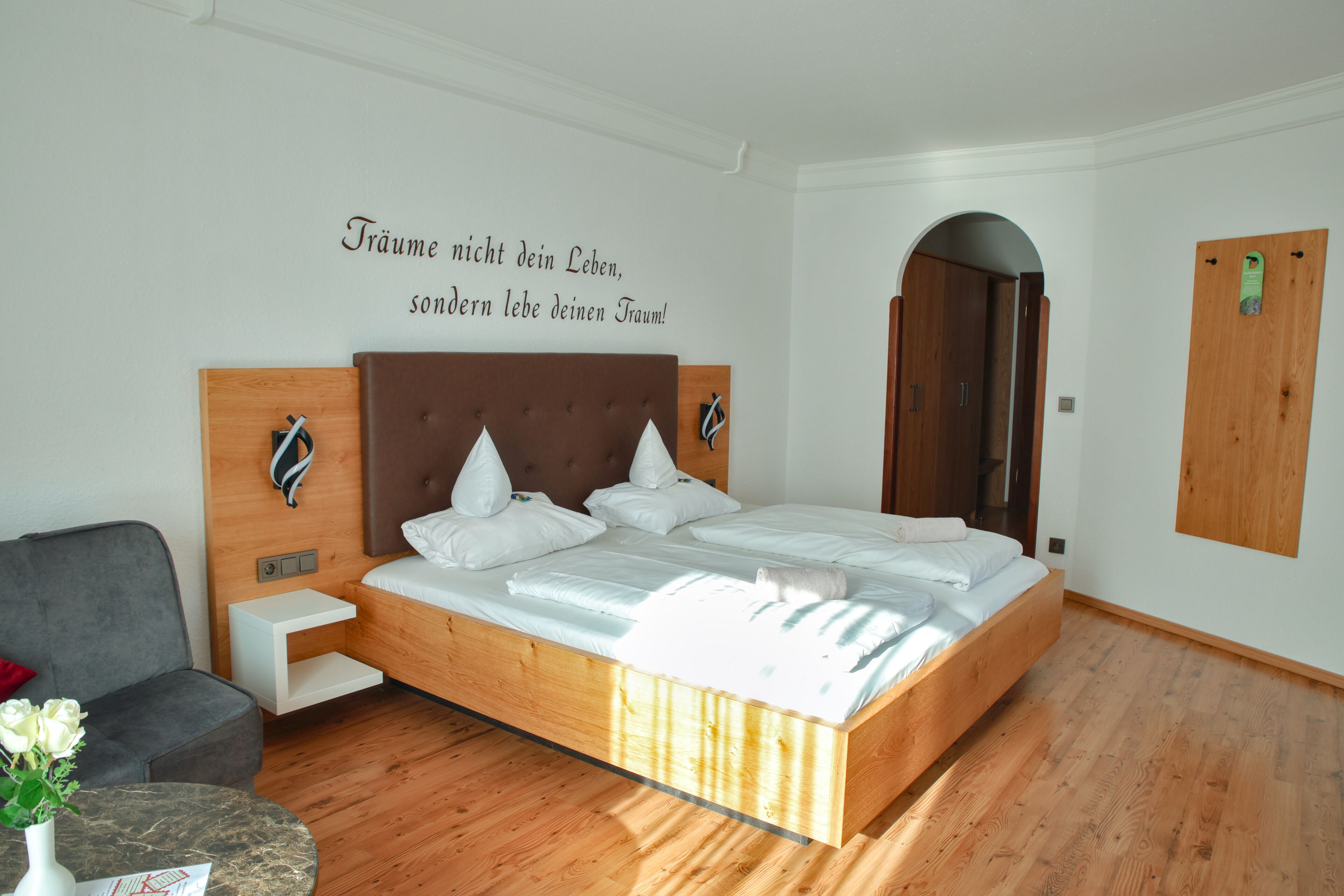 Un dormitorio moderno con una cama de madera, un sillón gris, una mesita con un florero y una pared con una cita en alemán.