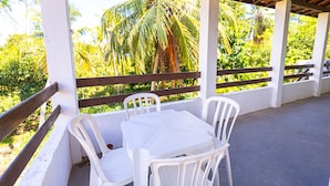 A covered outdoor seating area with white plastic chairs and a table, surrounded by white pillars and a wooden railing, overlooking a lush green landscape. - Casa com Piscina em Coqueiro de Arembepe (Camaçari)