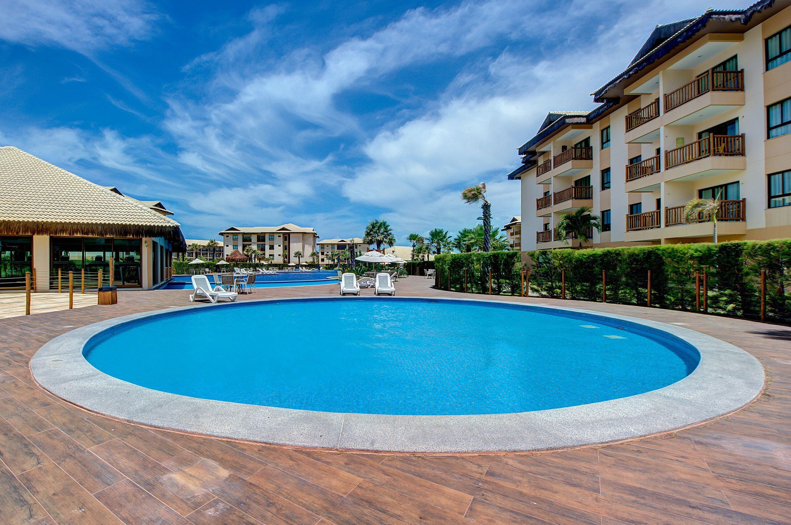 A large circular swimming pool with lounge chairs, surrounded by a wooden deck and a building with balconies.