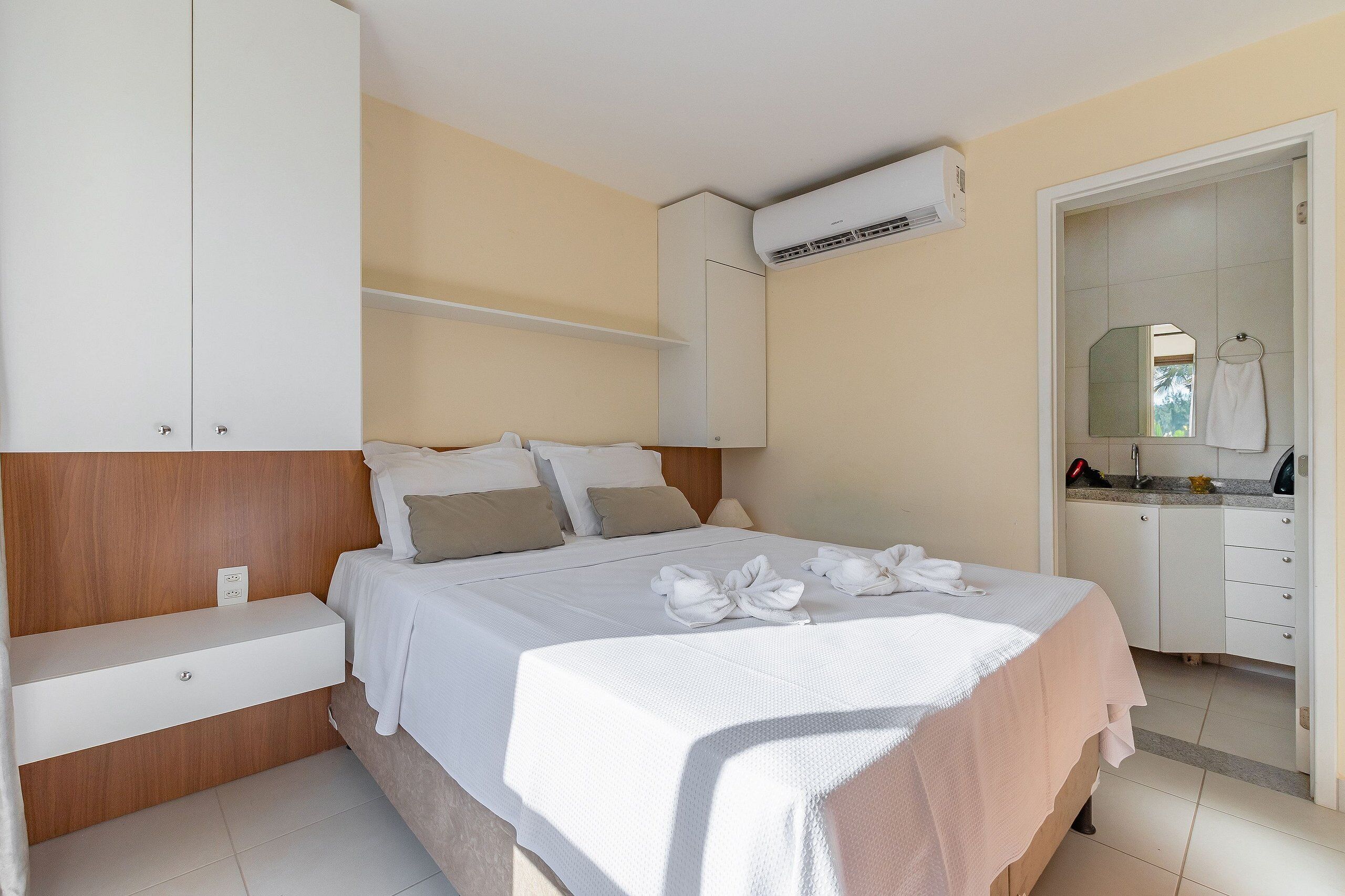Un dormitorio ordenado con cama, mesitas de noche, armario y baño visible a través de una puerta abierta.