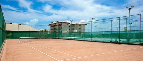 An outdoor tennis court with a green fence, a net, and a building in the background.