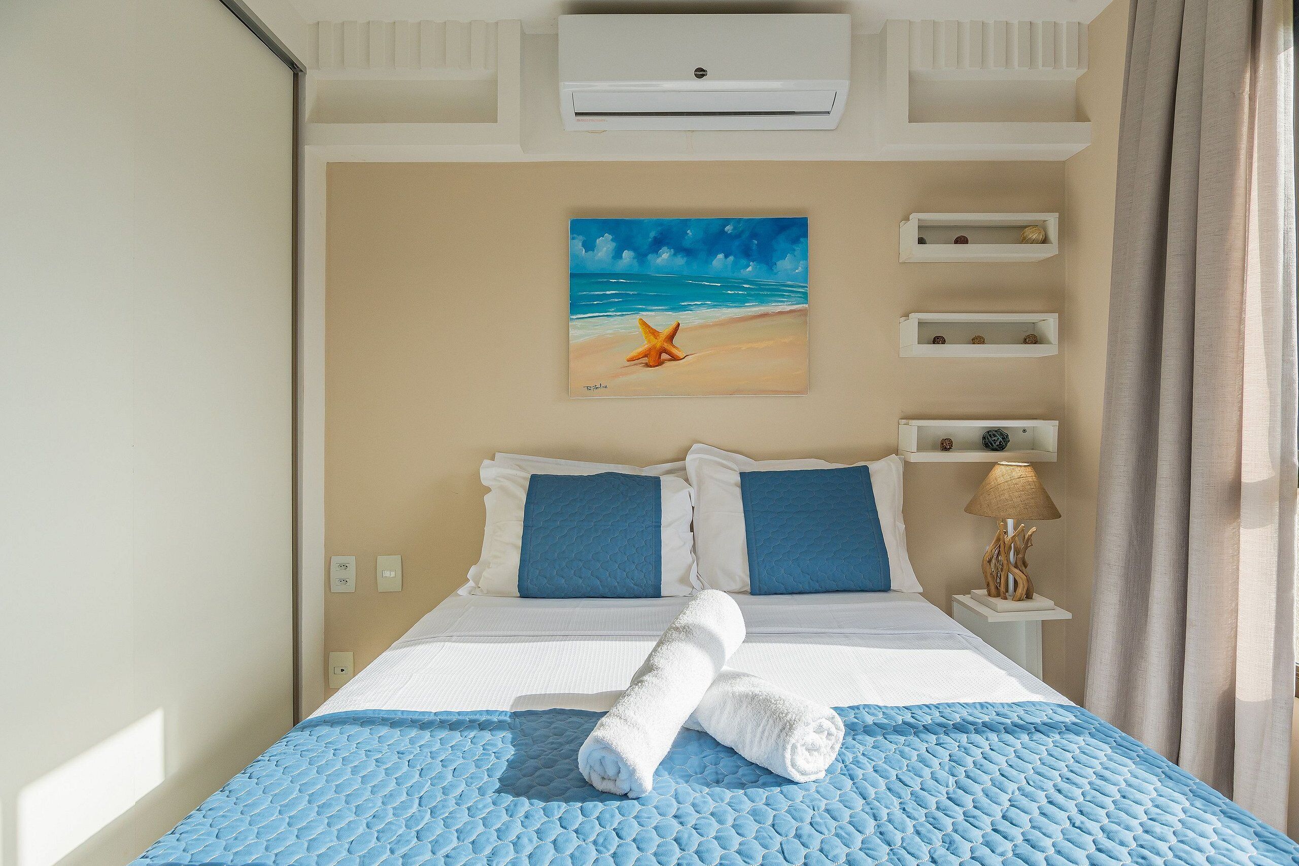 Quarto de hotel com uma cama, um quadro com uma cena de praia, uma mesa de cabeceira com abajur e uma janela com cortinas.