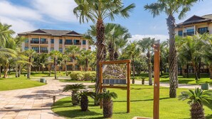 A courtyard with palm trees, a paved walkway, and a building with balconies. - VG Sun #310 - Quadruple Apartment by Carpediem (Caucaia)