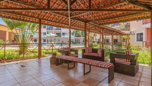 A covered outdoor seating area with a tiled floor, wicker furniture, and a view of a pool and buildings in the background. - Vila do Mar #112 - Terreo Apartment By Carpediem (Aquiraz)