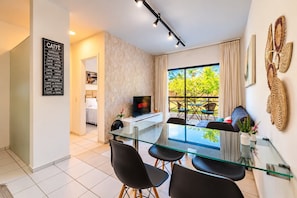 A modern living room with a glass dining table, black chairs, a flat-screen TV, and a balcony with a view of greenery. - Vila do Mar #112 - Terreo Apartment By Carpediem (Aquiraz)