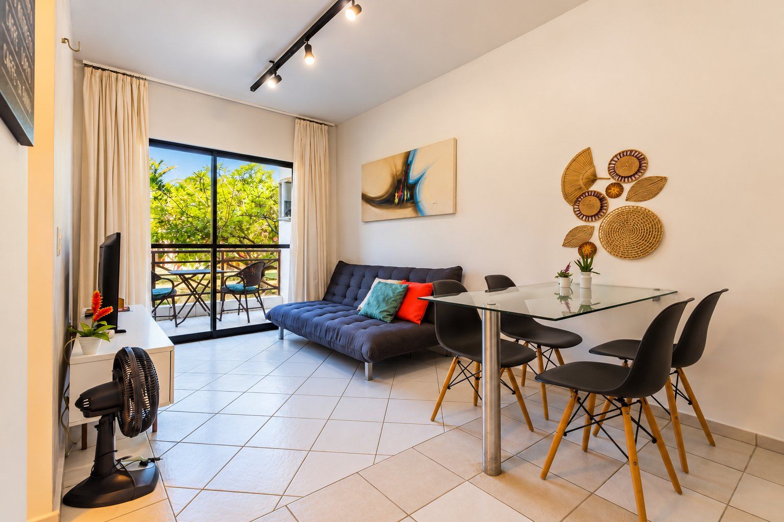 Uma sala de estar moderna com uma mesa de jantar de vidro, cadeiras pretas, um sofá com almofadas, uma TV de tela plana e uma grande janela com cortinas.