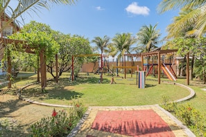 A playground with swings, slides, and climbing structures surrounded by palm trees and a brick pathway. - Vila do Mar #112 - Terreo Apartment By Carpediem (Aquiraz)