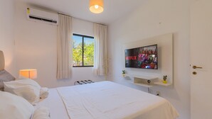 A hotel room with a bed, a TV mounted on the wall, a window with curtains, and a wall-mounted air conditioner. - Vila do Mar #112 - Terreo Apartment By Carpediem (Aquiraz)