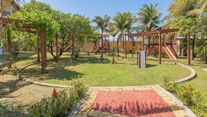 A playground with swings, slides, and climbing structures surrounded by palm trees and a brick pathway. - Vila do Mar #112 - Terreo Apartment By Carpediem (Aquiraz)
