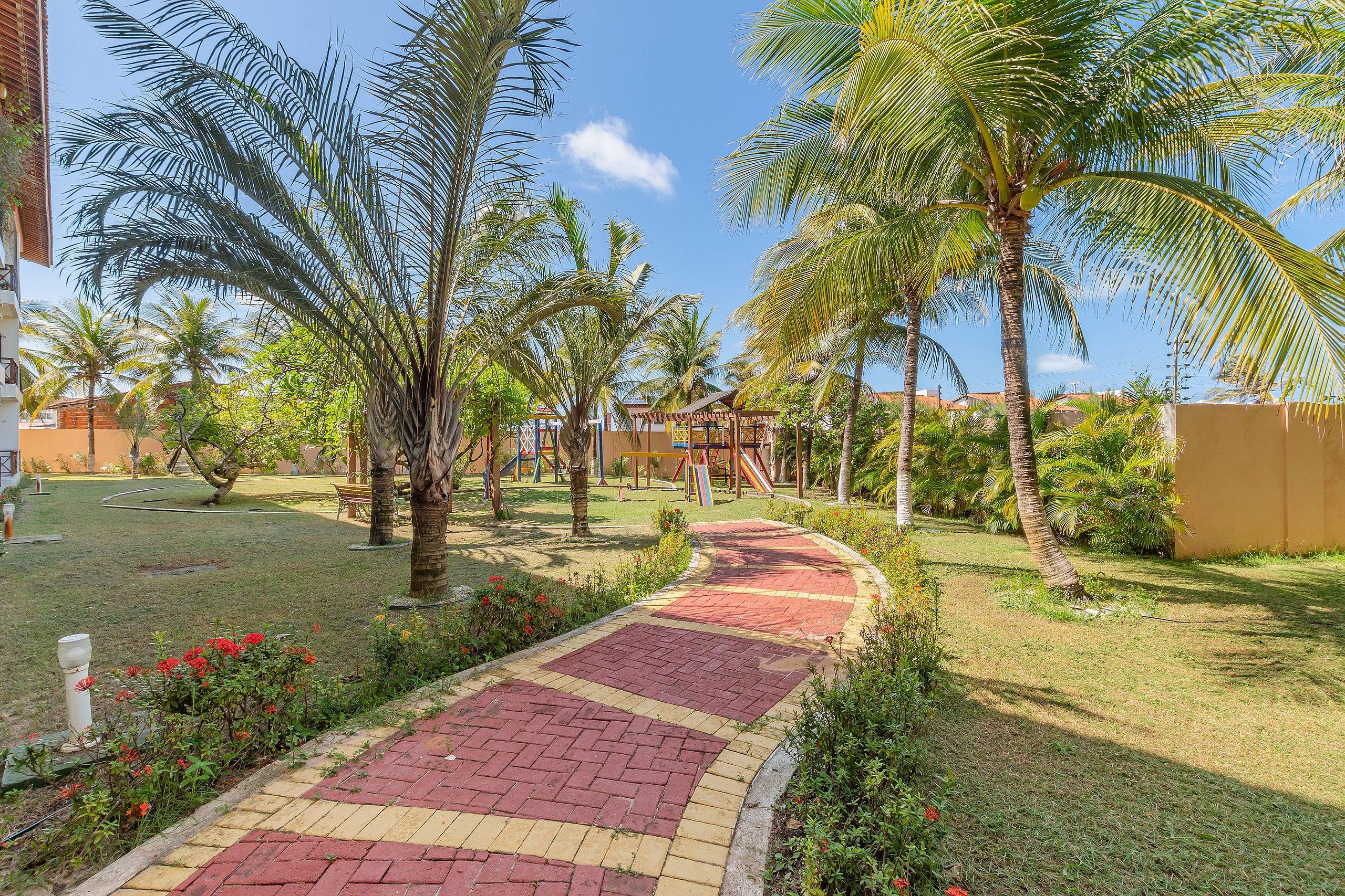Un sendero pavimentado que atraviesa un jardín con palmeras y un parque infantil al fondo.