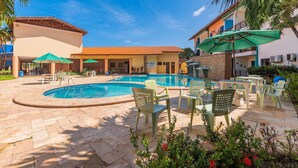 A swimming pool area with lounge chairs and umbrellas, surrounded by buildings with a tiled roof and a clear blue sky. - Vila do Mar #112 - Terreo Apartment By Carpediem (Aquiraz)