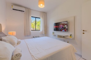 A hotel room with a bed, a TV mounted on the wall, a window with curtains, and a wall-mounted air conditioner. - Vila do Mar #112 - Terreo Apartment By Carpediem (Aquiraz)
