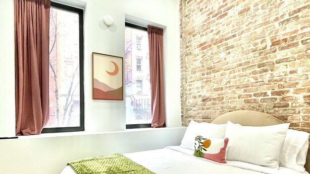 A hotel room with a brick wall, a large bed with white bedding, and two windows with curtains. - Hudson Yards Modern 1 bedroom (New York)