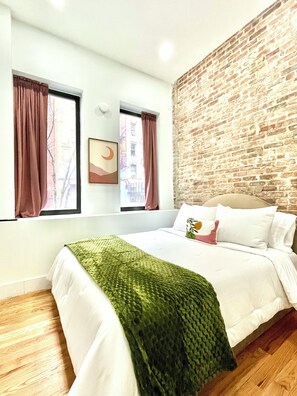 Habitación de hotel con pared de ladrillo, una cama grande con ropa blanca y dos ventanas con cortinas.