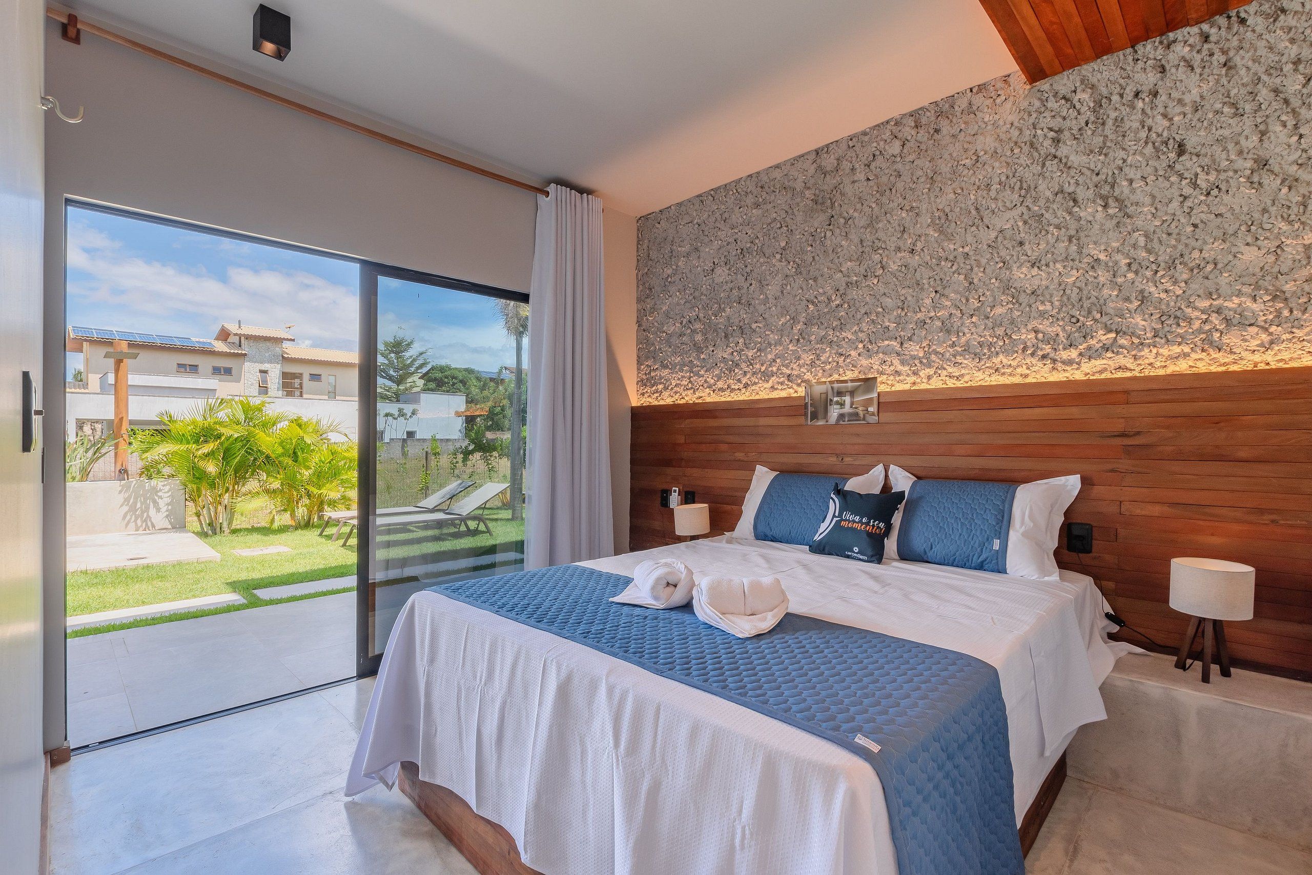 A bedroom with a large bed, a wooden headboard, a view of the outdoors, and a small table with a lamp.