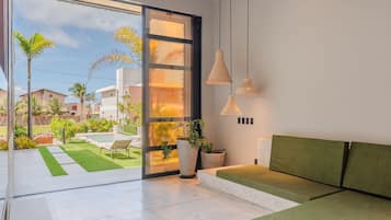 Un interior moderno con un ventanal corredero que da a un jardín, un sofá verde e iluminación colgante.