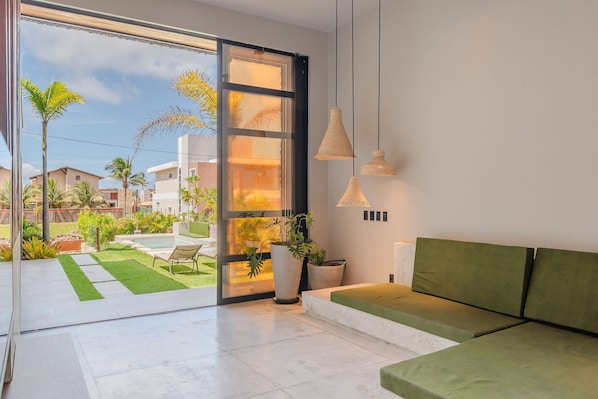 Un interior moderno con un ventanal corredero que da a un jardín, un sofá verde e iluminación colgante.