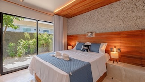 Ein modernes Hotelzimmer mit einem großen Bett, einem Holz-Kopfteil, einem kleinen Nachttisch mit Lampe und Blick auf Grünflächen.