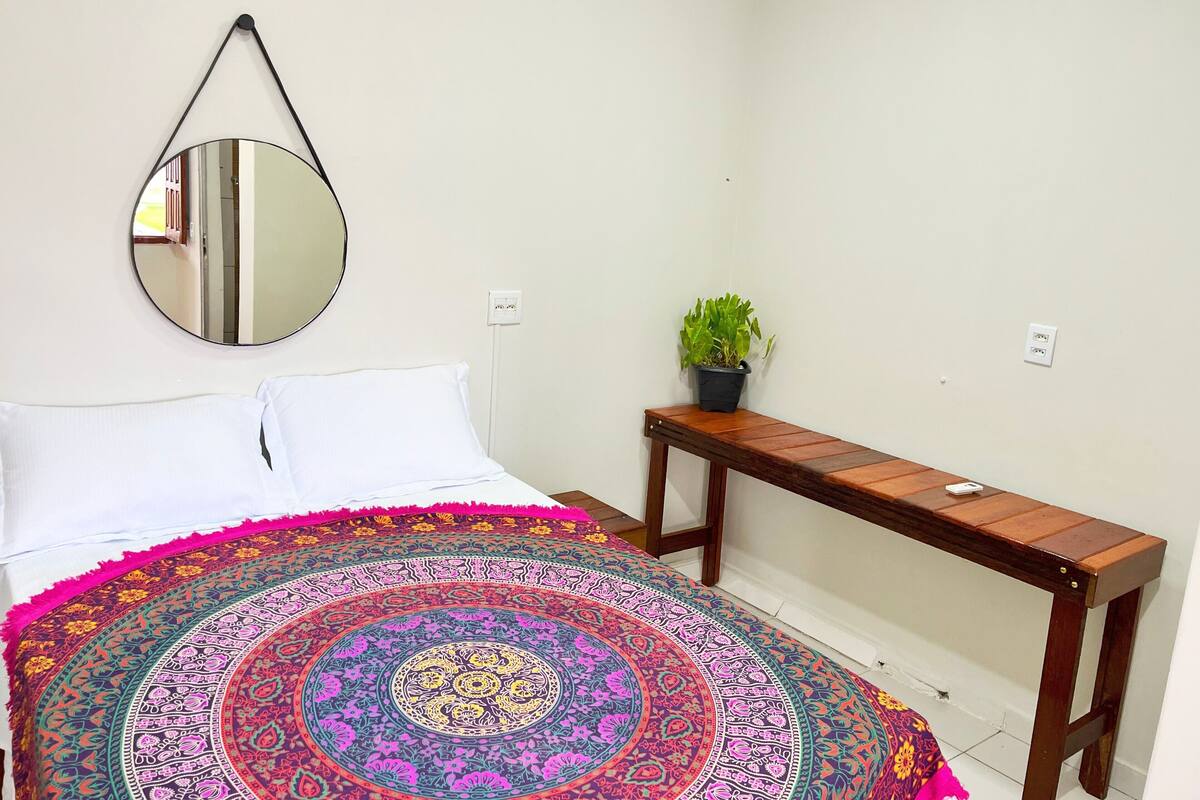 Une chambre à coucher avec un lit, un miroir rond, une table de chevet ornée d’une plante et une tapisserie aux couleurs vives.